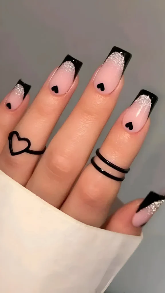 Long square nails with black tips, a sheer base featuring a silver glitter gradient fade from the tip, and a small black heart motif.