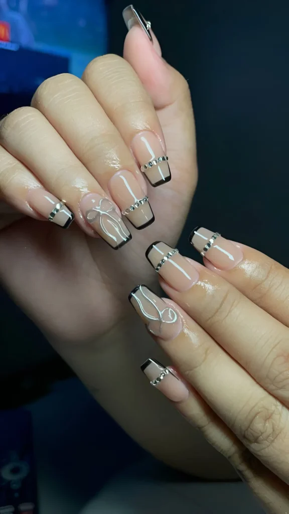 Square nails with sheer nude base, black French tips, a horizontal silver line with rhinestones, and silver line art bow accents.