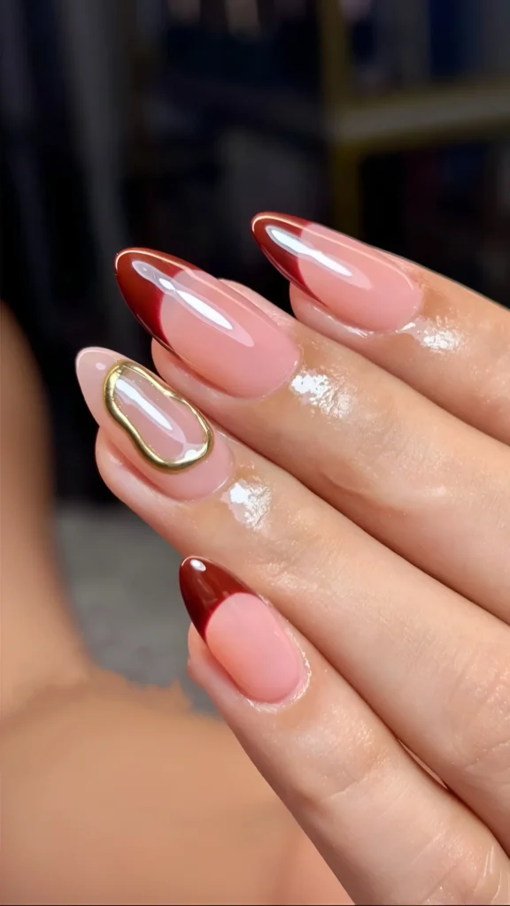 Long almond nails with deep red curved tips and an accent nail with an abstract gold metal embellishment.