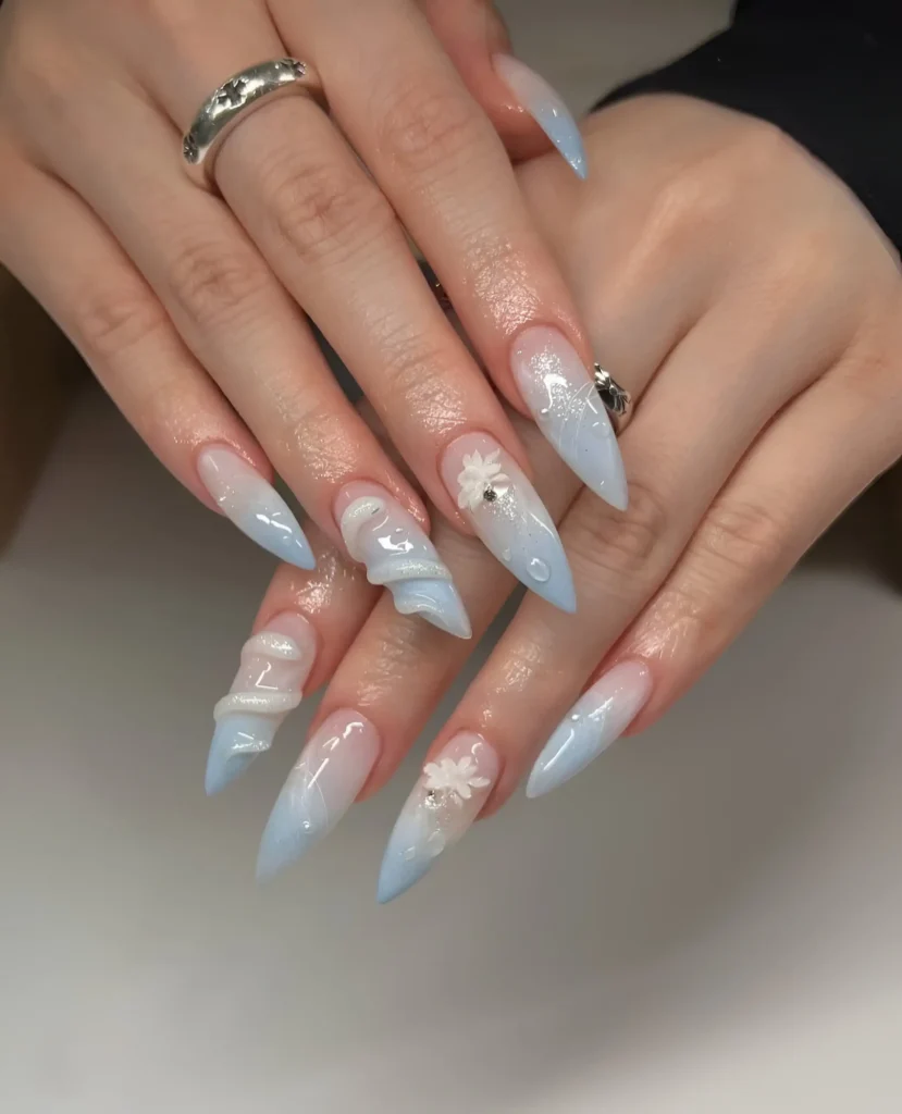 Long almond nails with sheer base, icy blue glitter gradient tips, white 3D ribbon swirls, and small white 3D flowers.