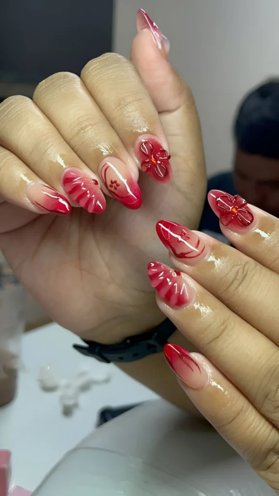 Pointed almond nails with a sheer nude to red ombré gradient, featuring dimensional red 3D sculpted flowers and ruffle textures on accent nails.