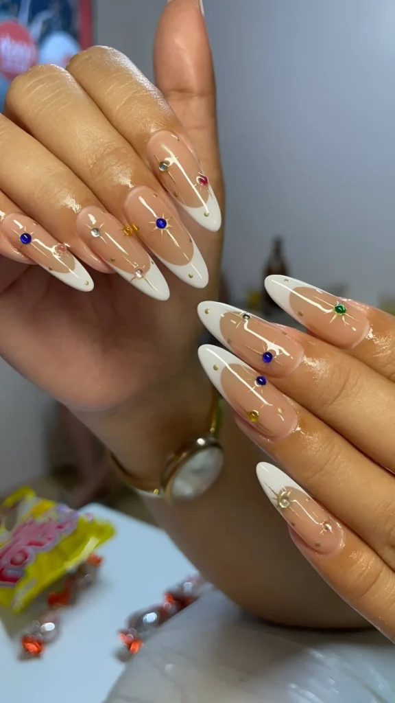 Almond nails with sheer nude base, classic white French tips, and colorful rhinestone accents (blue, green, yellow) with small gold studs.