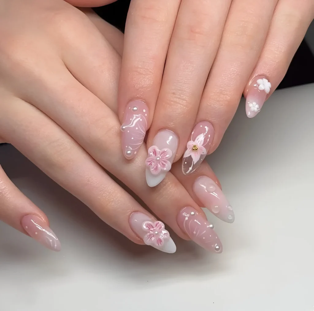 Long almond nails with sheer milky base featuring delicate pink and white 3D flowers, fine white painted details, and scattered pearl accents.