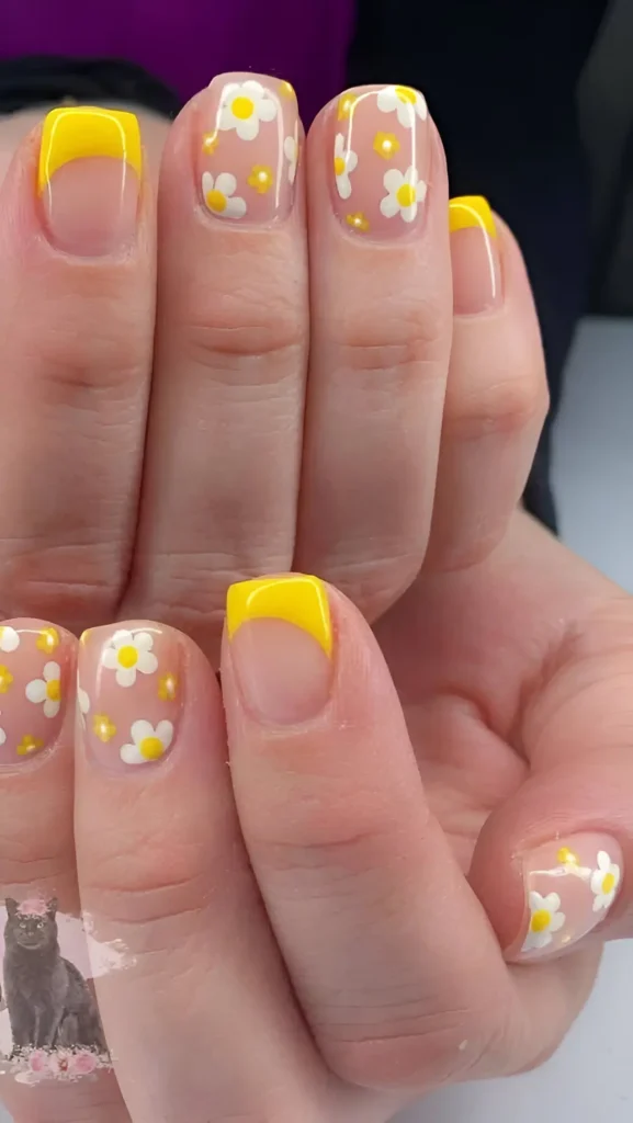 Short square nails showing a mix of bright yellow classic French tips and nails with a sheer base covered in a pattern of small white daisies with yellow centers.