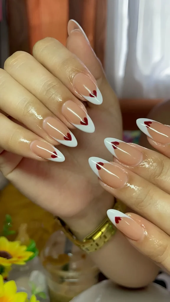 Almond nails with sheer nude base featuring deep white V-shaped French tips accented with a small red heart at the point.