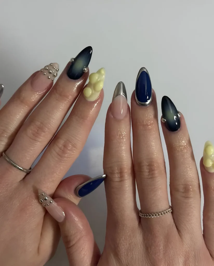 Almond nails mixing deep blue polish with silver accents, pale yellow 3D texture, silver studded nails, and metallic silver tips.