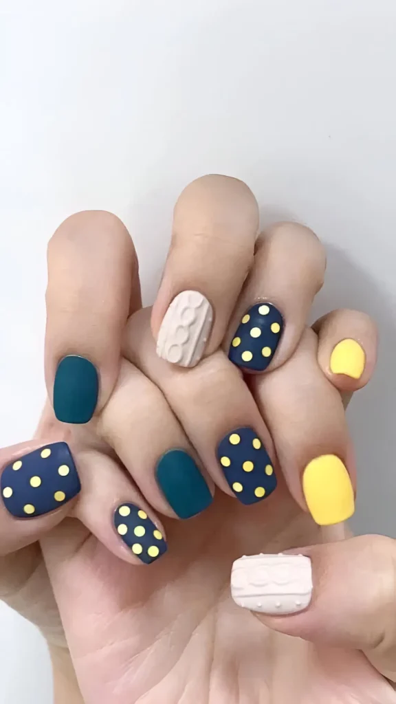 Short square matte nails: solid pastel yellow, dark teal with yellow polka dots, and off-white nails with a raised 3D cable-knit sweater texture.