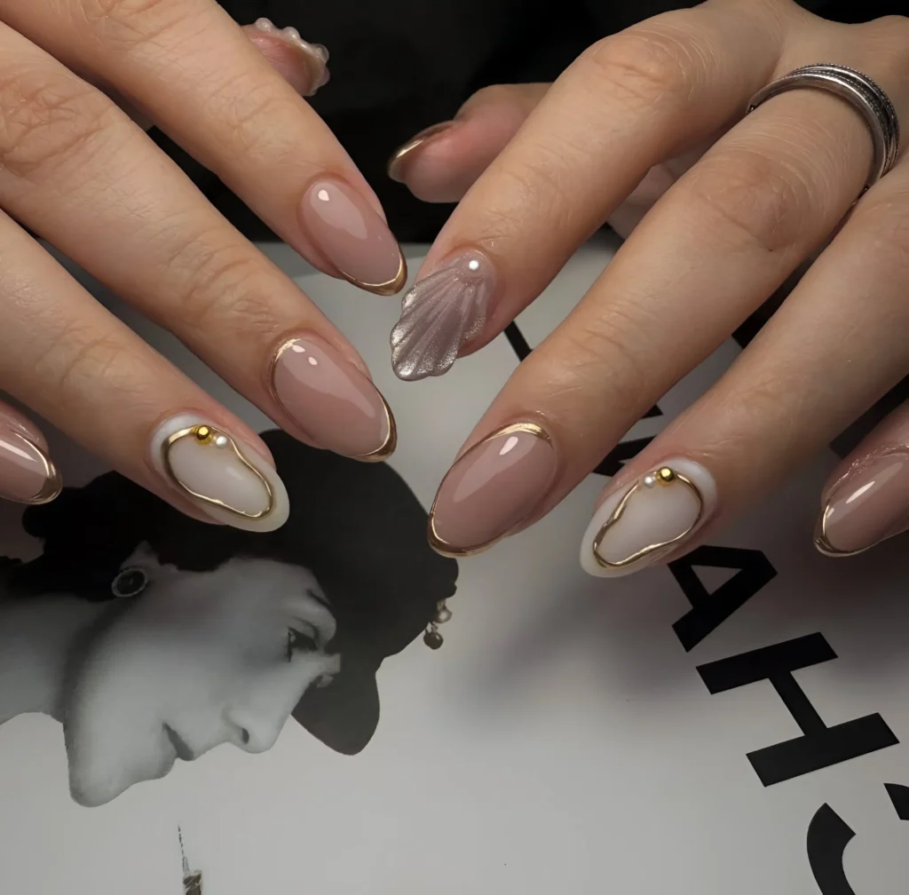 Chic nude almond nails with gold outlines, abstract white shapes, and subtle single pearl accents.