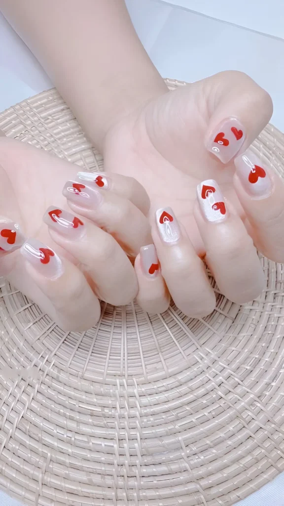Sweet minimalist look: scattered tiny red heart motifs on a sheer milky base on short squoval nails. A simple classy nail design.