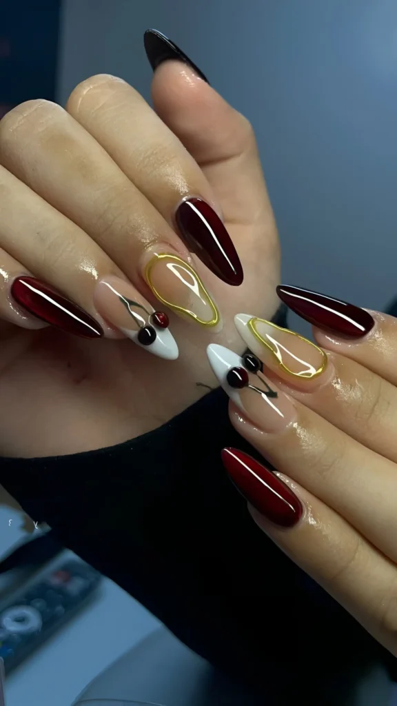 Stiletto nails mixing solid oxblood red with sheer nude bases featuring abstract gold outlines and red, white, and black cherry-like motifs.