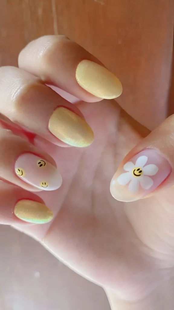 Almond-shaped nails featuring a mix of pastel yellow shimmer polish, sheer nails with scattered small yellow smiley faces, and a sheer nail with a white daisy having a yellow smiley center.