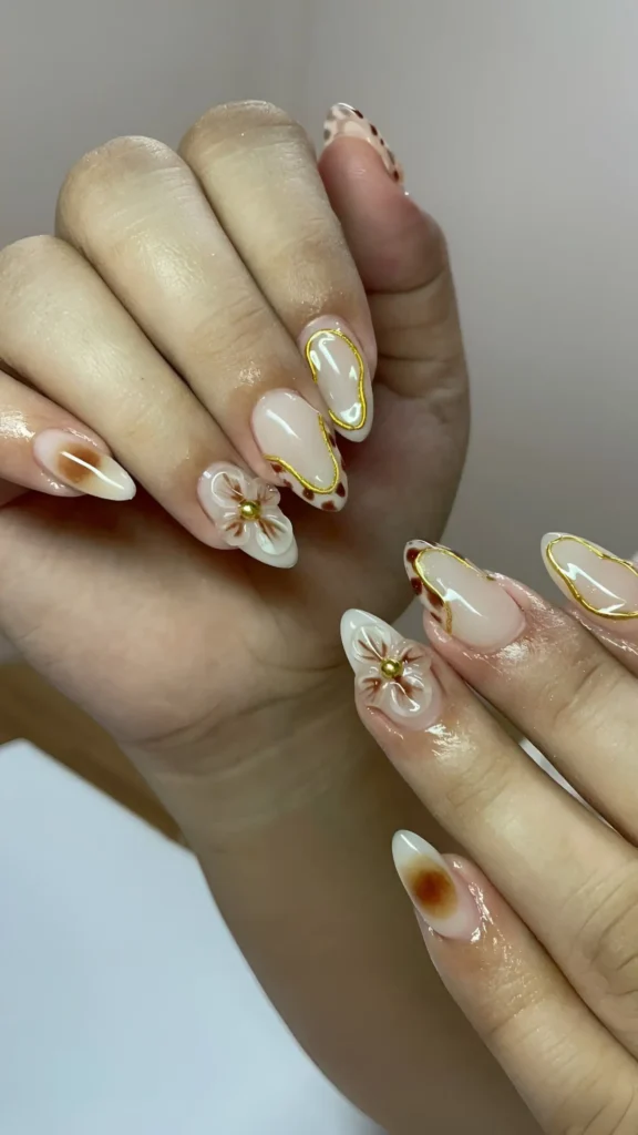 Almond nails with sheer nude base featuring tortoiseshell accents, dimensional cream 3D flowers with brown centers, and abstract gold outlines.