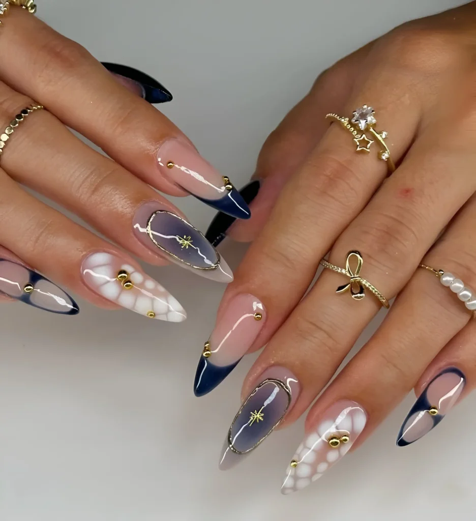 Elegant stiletto nails mixing navy V-French tips, textured white blooms, and sheer wash framed in gold with celestial accents.