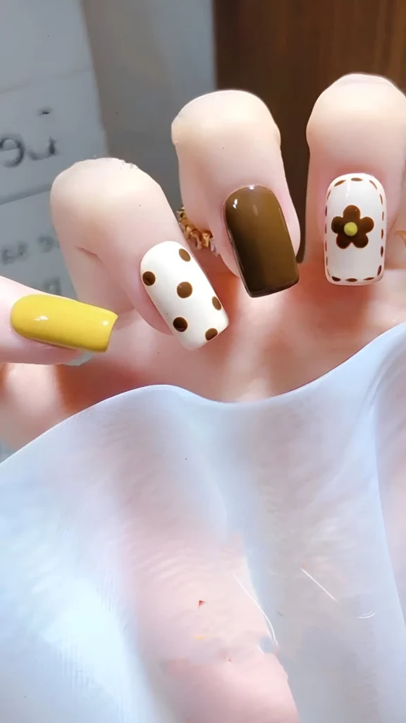 Square nails with a mix-and-match design: solid mustard yellow, solid brown, white with brown polka dots, and white with a brown flower outline having a yellow center.