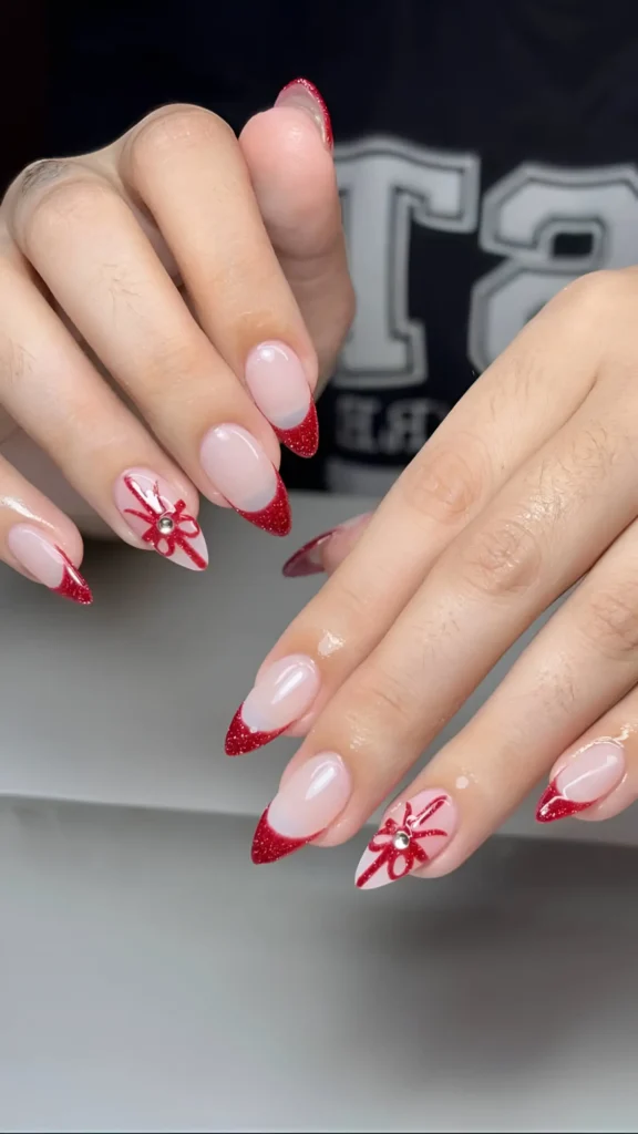 Pointed almond nails with sheer base, red glitter V-shaped tips, and accent nails with red bow art and gem centers.