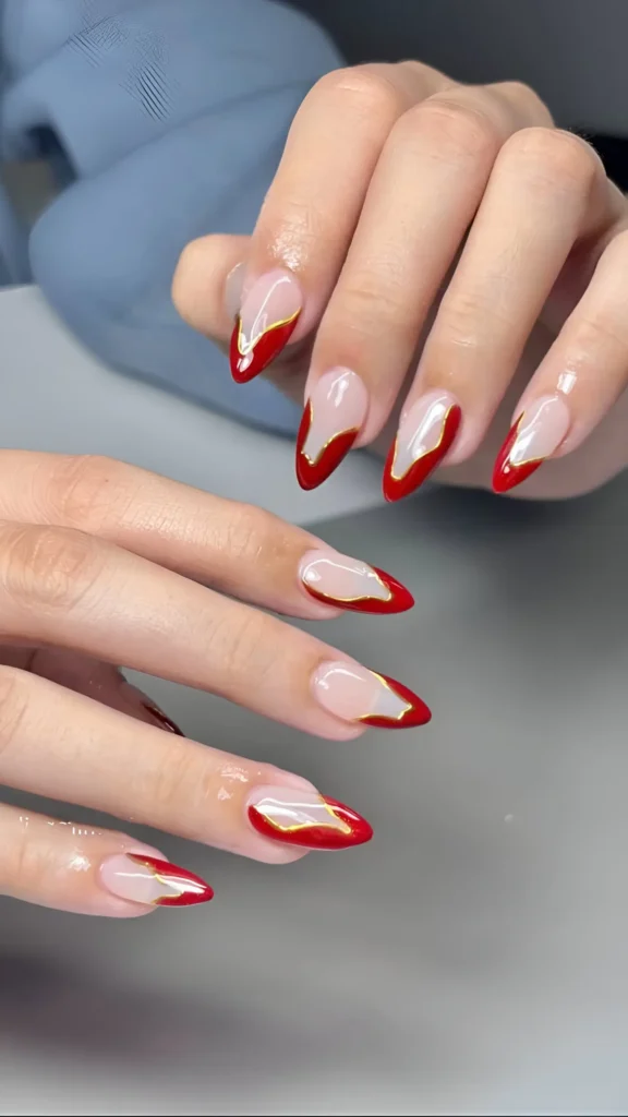 Pointed almond nails with sheer base, abstract bright red tips outlined in metallic gold.