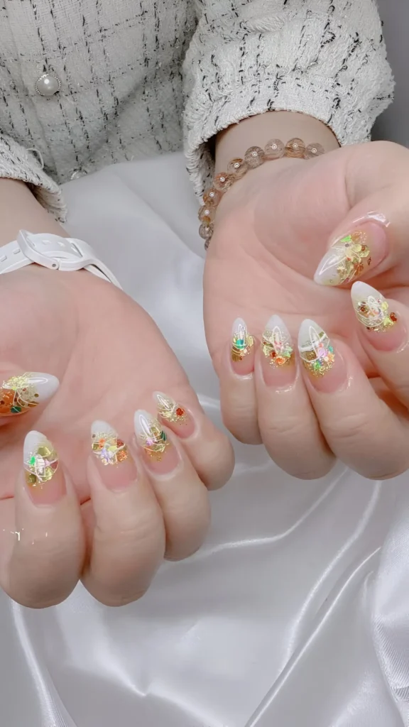 Opulent almond nails with milky white base decorated with clusters of gold foil and colorful micro-rhinestones near cuticles.