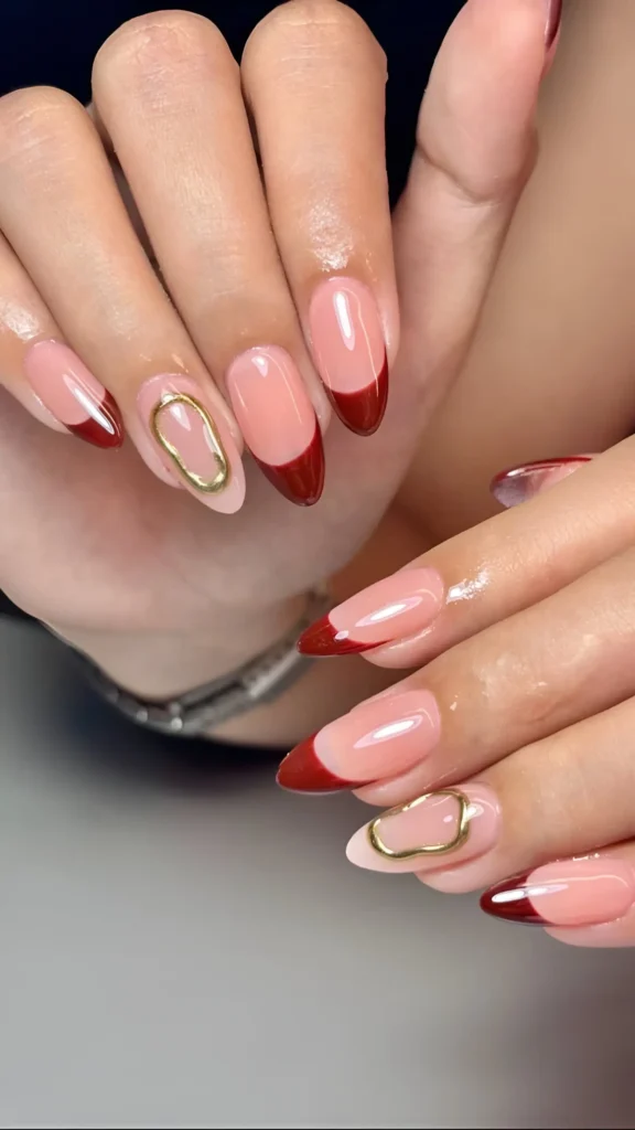 Almond nails with sheer base, deep earthy red curved tips, and accent nails featuring abstract gold hardware.