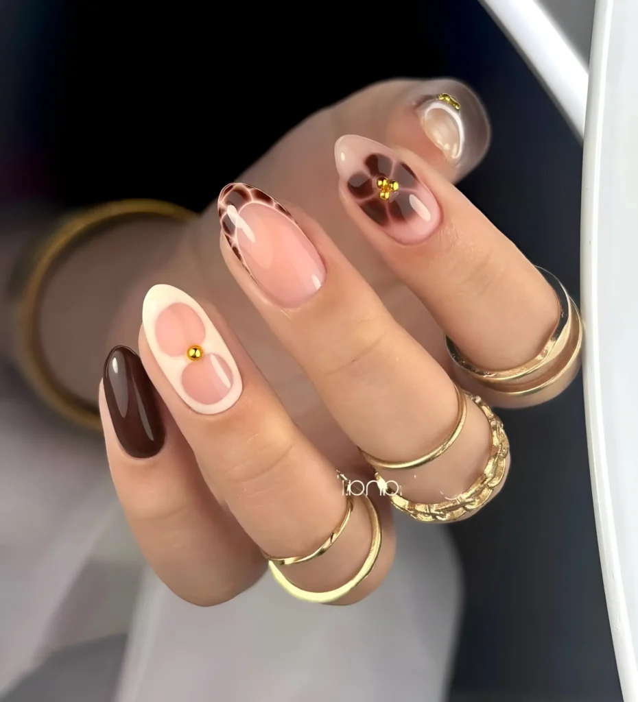 Sophisticated almond nails mixing solid chocolate brown, cream accents, abstract florals, and minimalist gold studs.