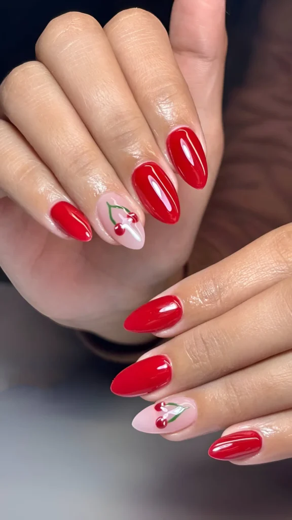 Glossy cherry red almond nails with one accent nail displaying cherry illustration on a sheer base.
