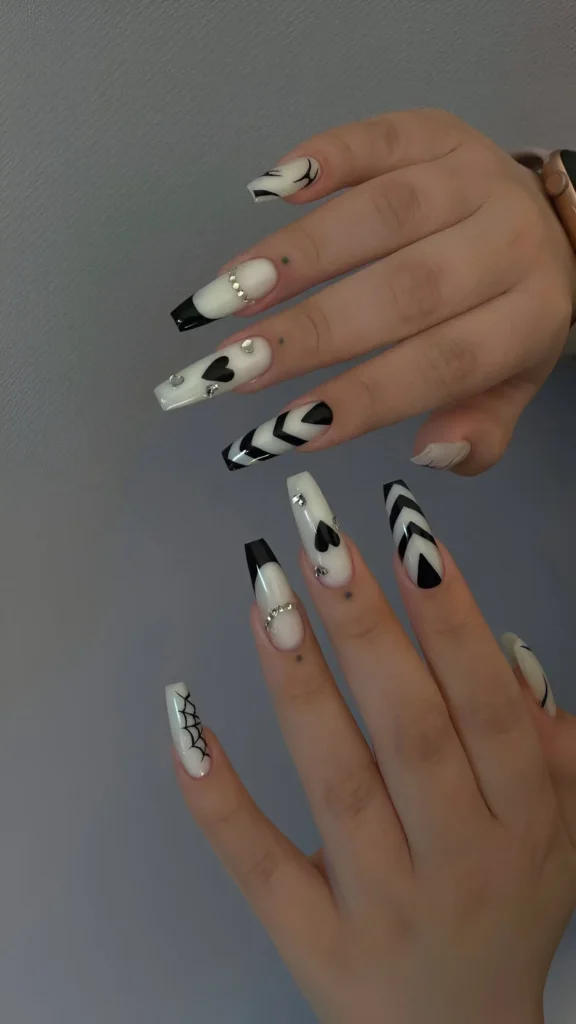 Long coffin nails with a milky white base featuring a variety of black graphic designs (hearts, chevrons, flames, web, French tips) and rhinestone accents.