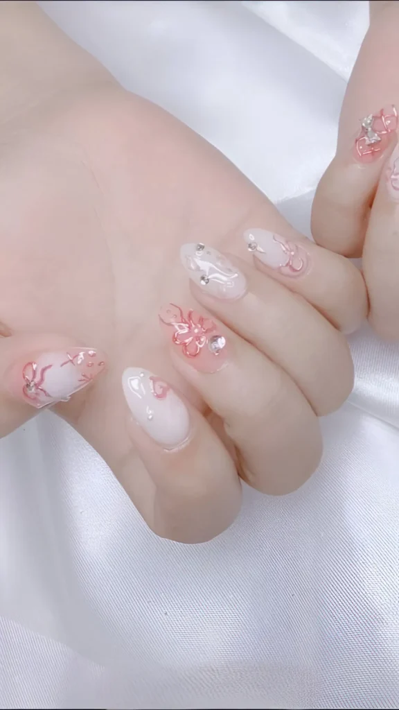 Ethereal almond nails: milky white base featuring delicately sculpted 3D bows and swirls in translucent pink gel with rhinestones.