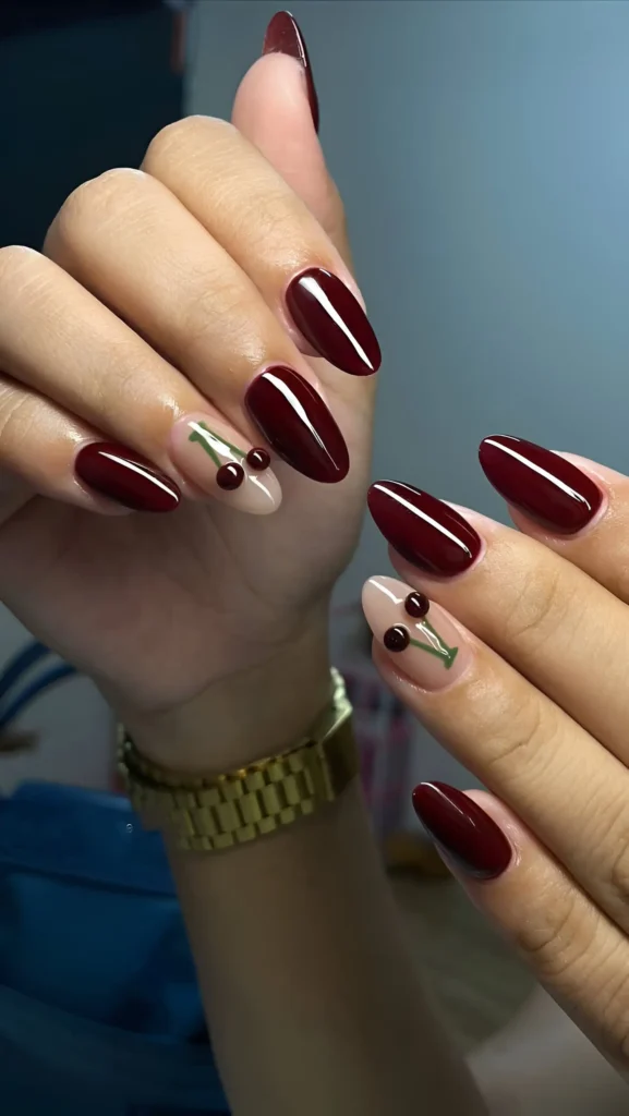 Almond nails mixing solid oxblood red polish with sheer nude accent nails featuring a painted red cherry motif with green stems.