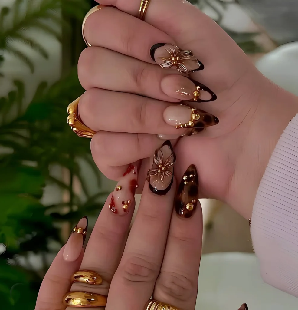 Elegant long nails combining translucent 3D florals, tortoiseshell patterns, dark V-tips, and gold stud accents.