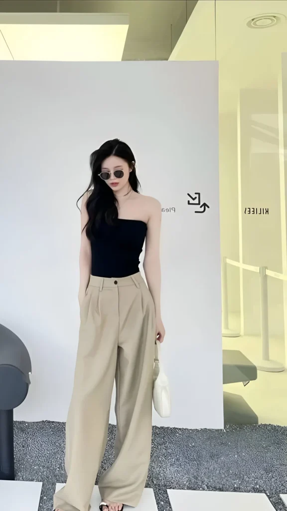 Woman wearing a black strapless tube top, beige high-waisted wide-leg trousers, and sunglasses, carrying a white handbag indoors