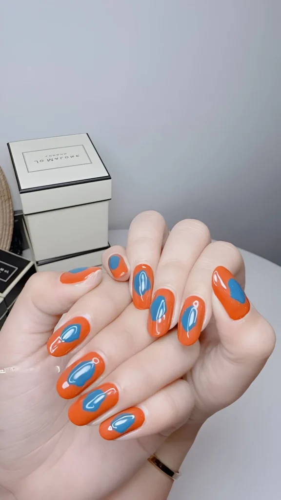 Modern abstract nail art: vibrant orange oval nails centered with a contrasting medium blue organic blob shape.