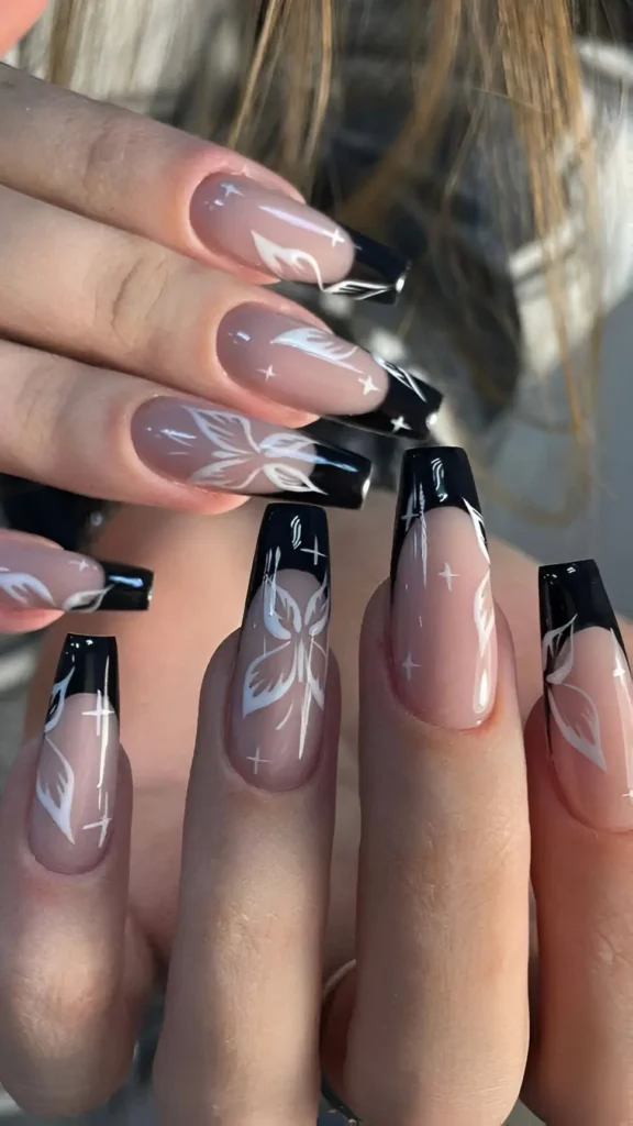 Long coffin nails with a muted taupe base, black French tips, and soft white stylized floral/leafy graphics and starburst accents.
