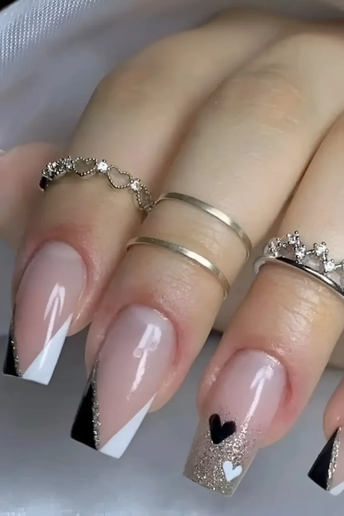 Square nails with sheer base featuring angled black and white color block tips divided by gold glitter, and a glitter accent nail with black and white hearts.