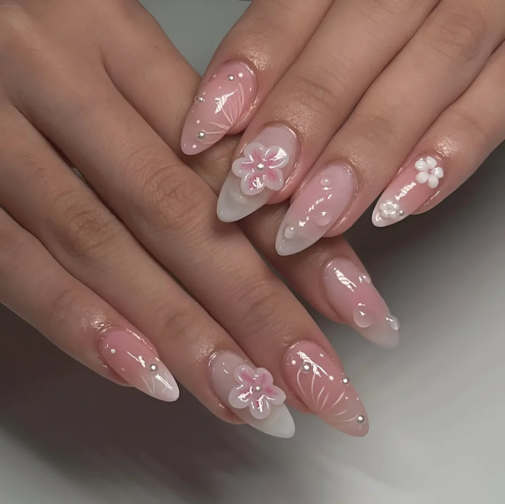 Almond nails with sheer pink/white ombré base featuring delicate pink and white 3D flowers, fine white linework details, and pearl/stud accents.