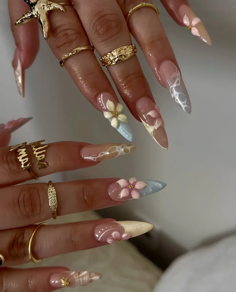 Long almond nails mixing muted 3D florals (pink, yellow), varied tips (grey marble, blue, yellow), white abstract lines, and gold accents on sheer base.