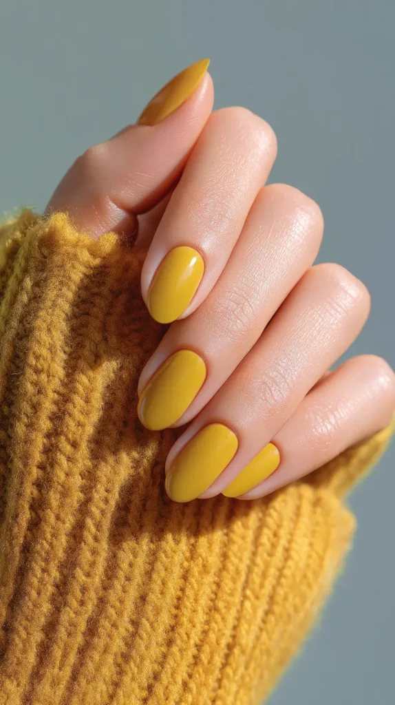 Close-up of manicured rounded nails in solid mustard yellow glossy polish, resting on a cozy matching knit sweater - showcasing chic yellow nail ideas.