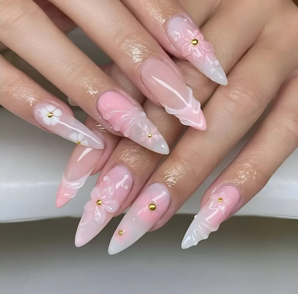 Long almond nails with sheer/white ombré base featuring pink and white 3D flowers, sculpted white textures, and gold bead accents.