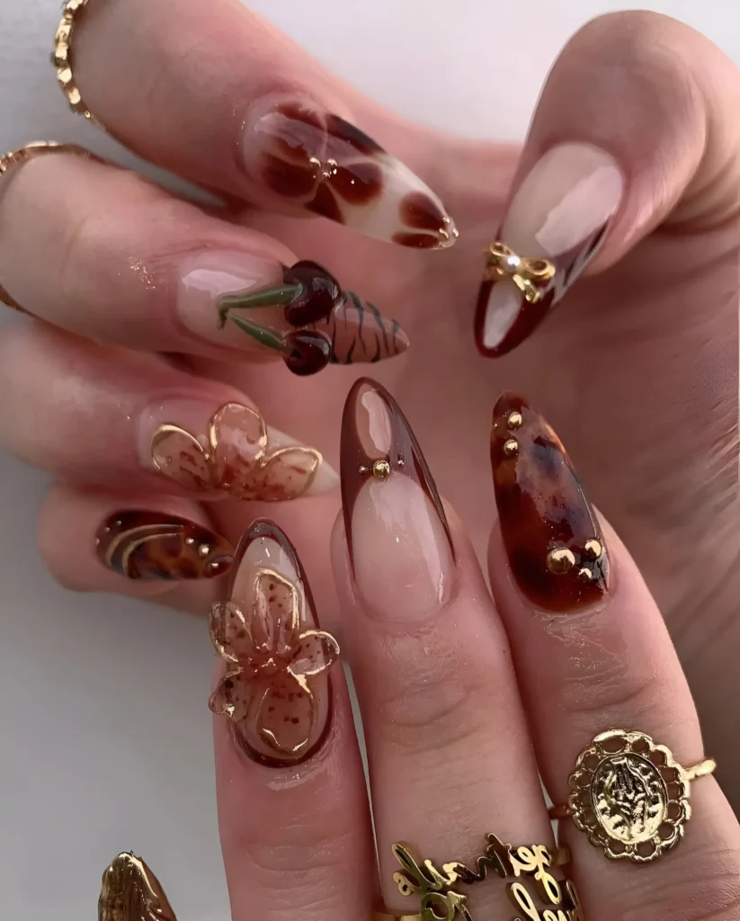 Chic pointed nails combining tortoiseshell patterns, translucent 3D florals, sculpted cherries, and gold stud accents.