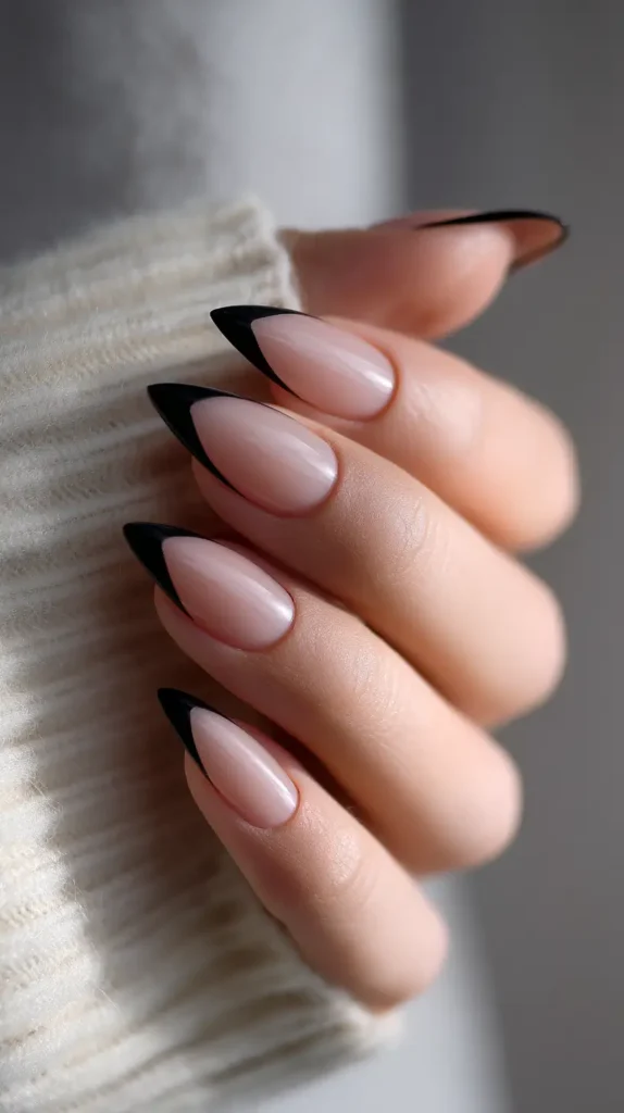 Long pointed almond nails with a sheer nude base featuring deep, sharp black V-shaped French tips.