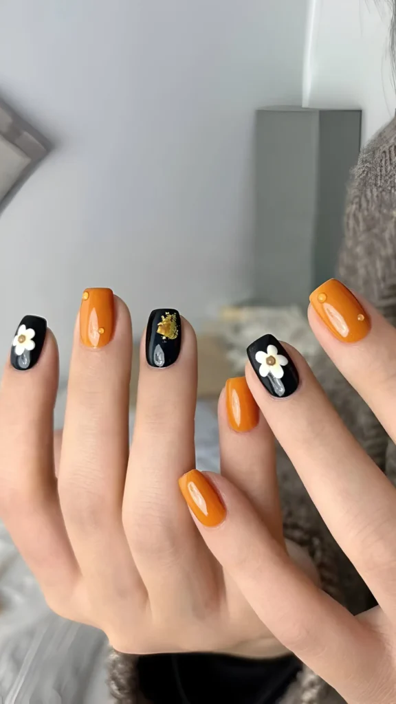 Hand showing a manicure alternating between glossy black nails with white flowers and solid warm orange nails.
