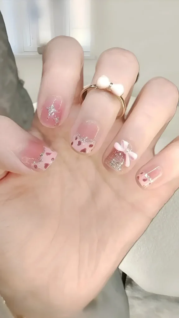 Cute pink nail design with tiny pink hearts, silver glitter stars, 3D pink bow, and rhinestone accents.