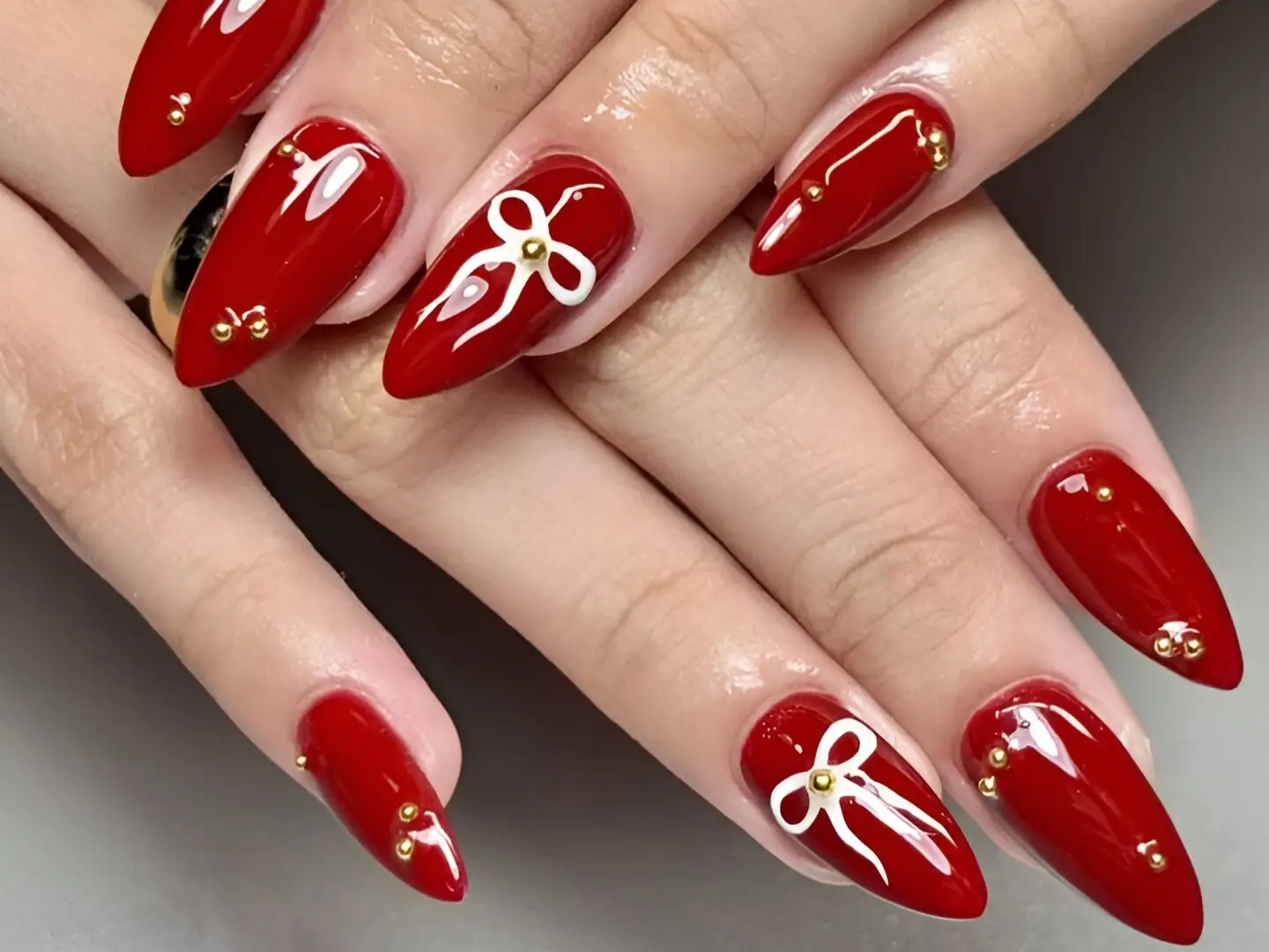 Red nail ideas featuring deep red almond nails decorated with white bow outlines, gold studs, and scattered gold accents.