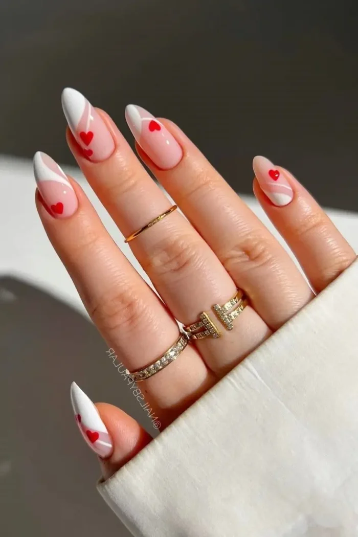 Close-up of almond-shaped nails featuring a modern French manicure with white swirls and small red heart details on a sheer pink base.