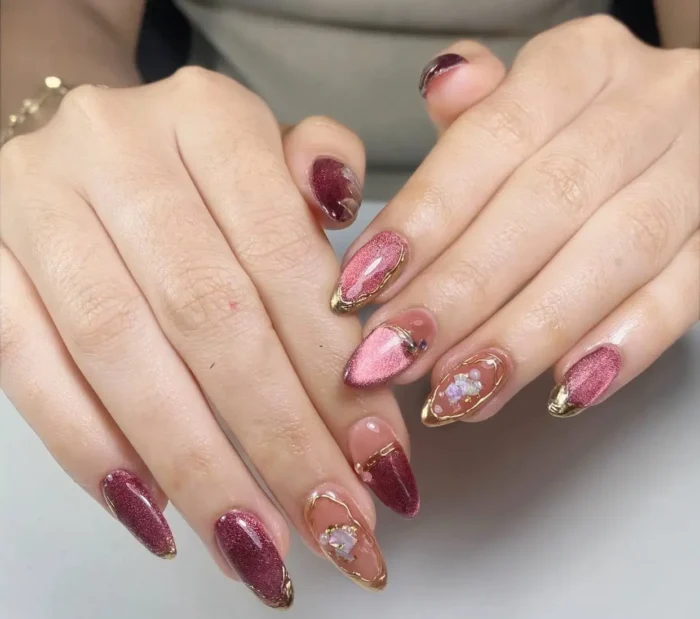 Almond nails with a mix of designs including glittery burgundy, pink cat-eye effect, and negative space with gold outlines and embellishments.