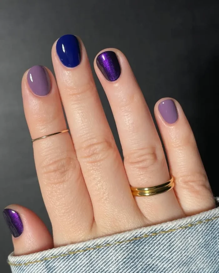 Simple gel manicure with mixed jewel tone colors including dusty purple, royal blue, and metallic purple on short nails.