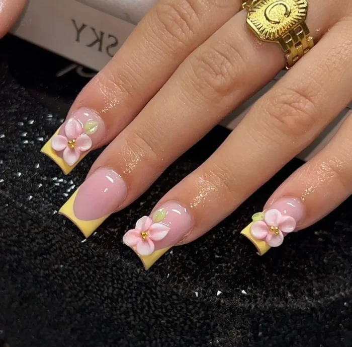 Square-shaped nails with pastel yellow French tips, adorned with 3D pink flower and subtle green leaf accents at the smile line.
