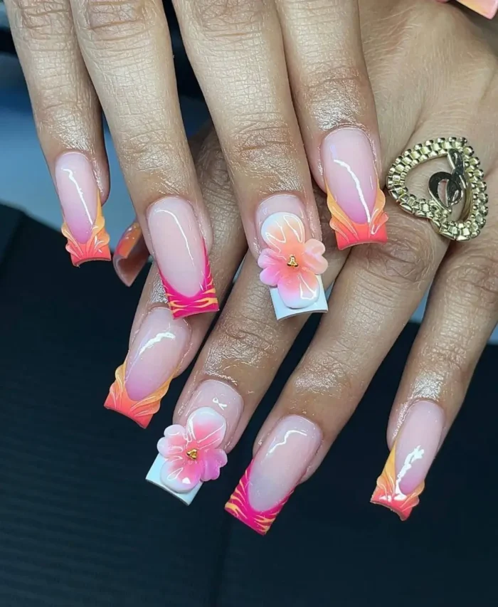 Bold summer manicure featuring fiery sunset ombré sculpted tips and 3D tropical flower accents on long square nails.