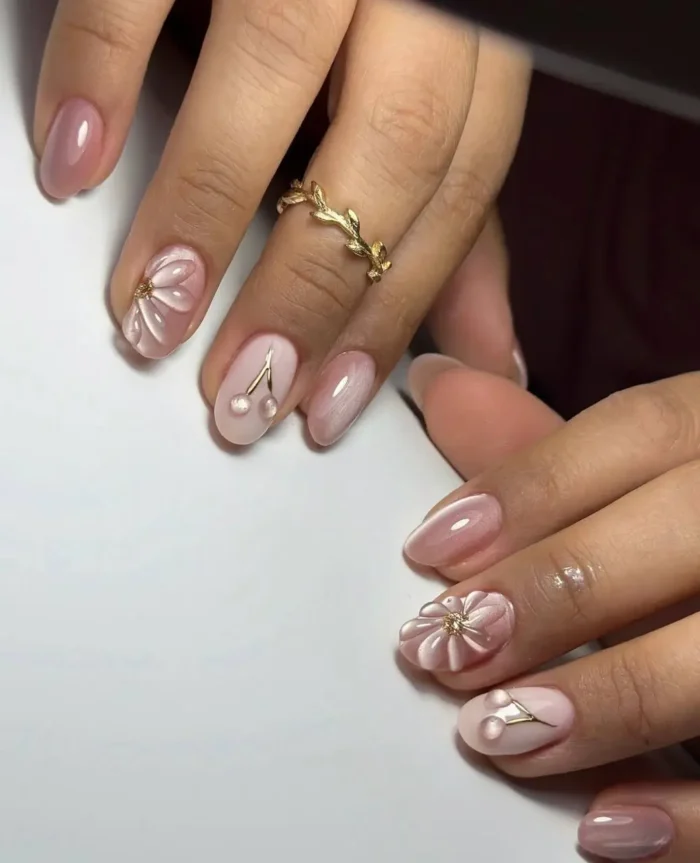 Short oval nails with a pearly pink base featuring delicate 3D white floral designs and minimalist gold charm accents.