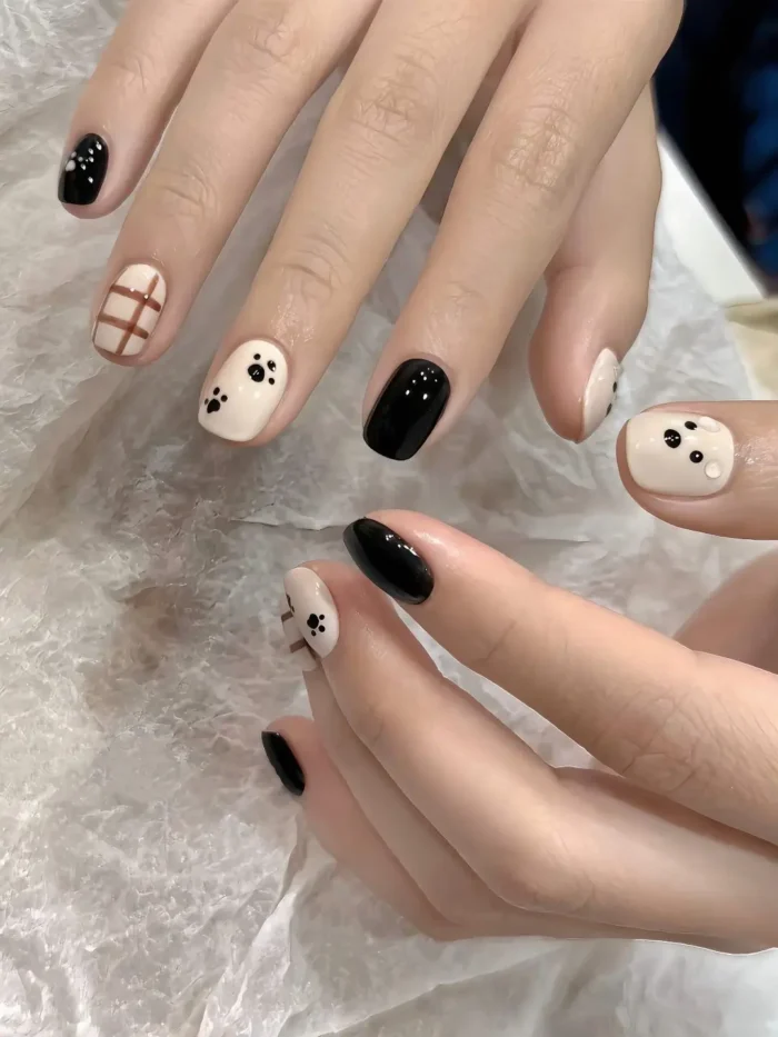 Short rounded nails with a mixed design featuring solid black polish, white nails with black paw prints, and a white nail with a brown grid pattern.