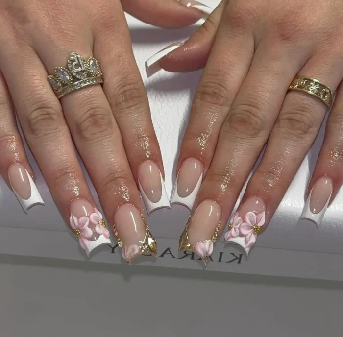 Glamorous summer French manicure with white tips, 3D pink flowers, and gold cuticle accents on long nails.