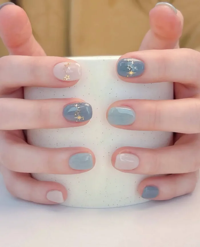 Short rounded nails with a mix of muted dusty blue, grey-blue, and sheer pink polish, accented with tiny gold star decals.